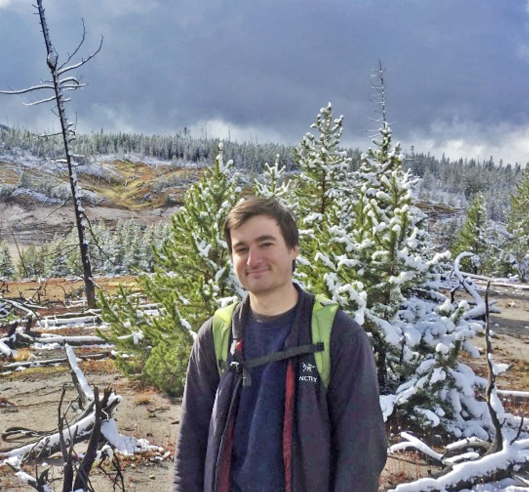 Image of Anthony Kohtz. He is smiling at the camera. There is a forest in the background and there is snow on a pine tree.