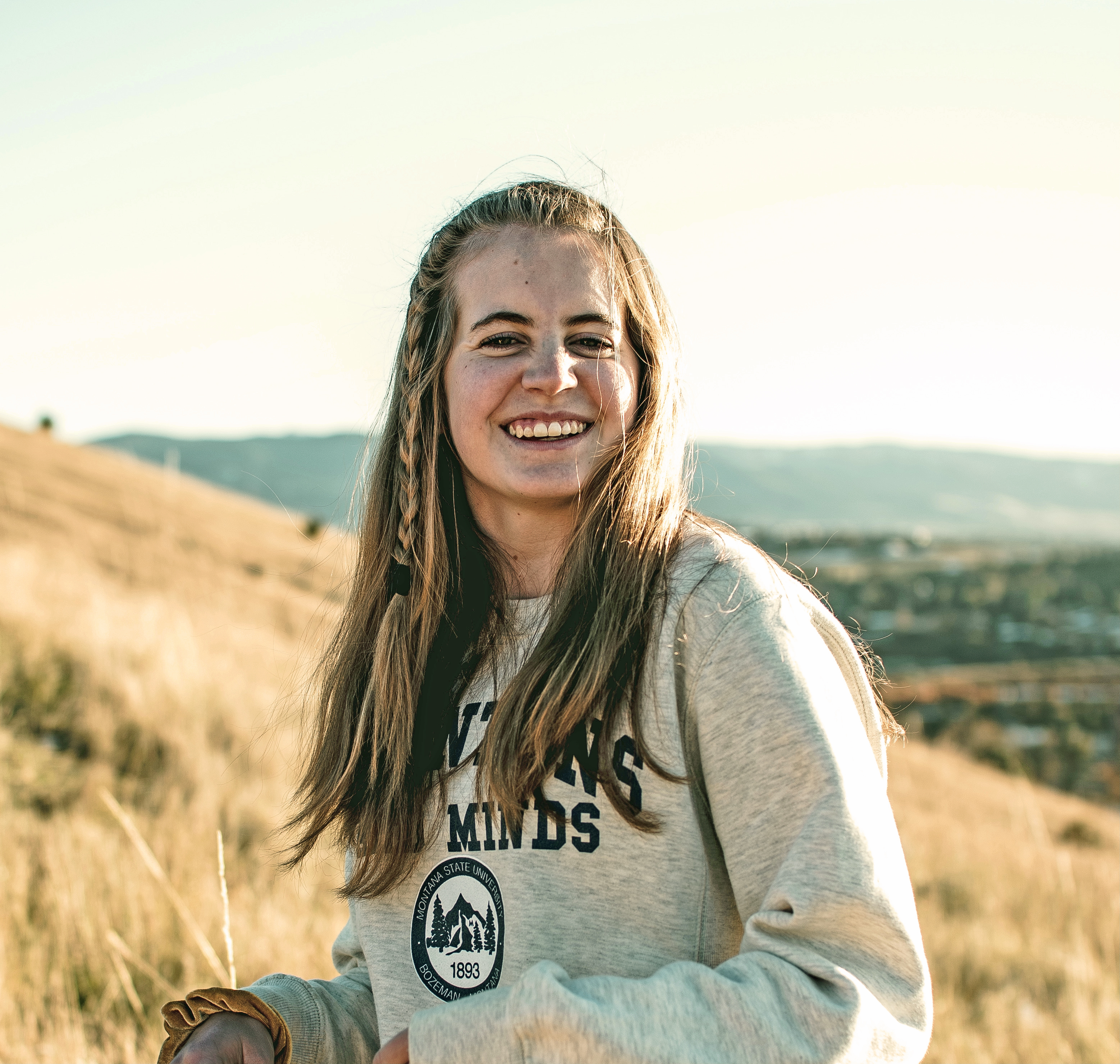 Image of Paige Schlegel smiling at the camera. In the background is a blurred hill. The grass is yellowing and a town is in the background.
